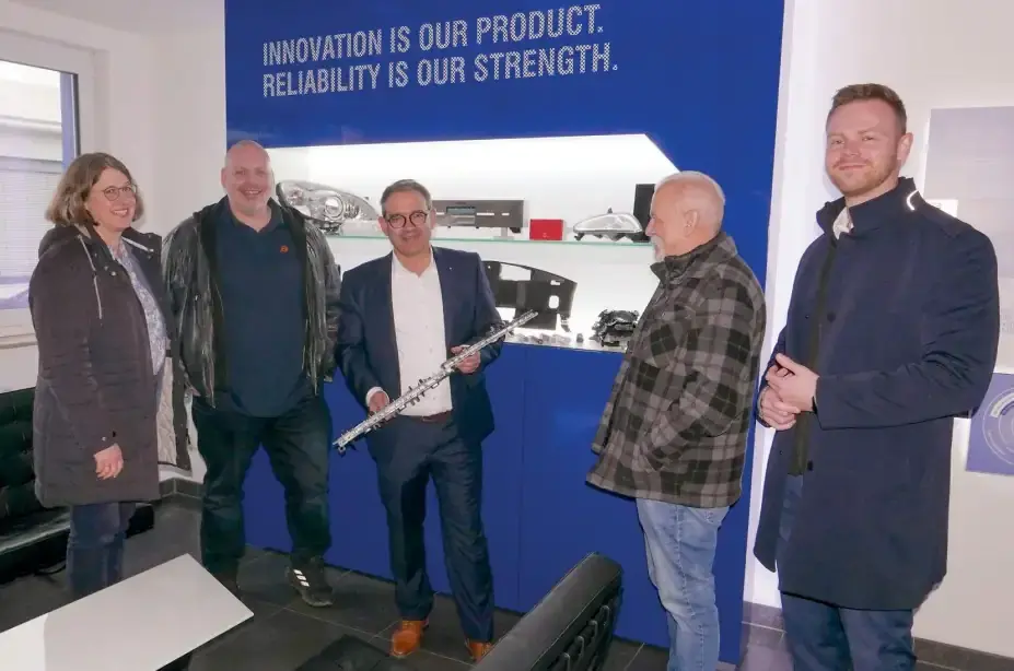Executives and visitors of Arnstädter Werkzeug- und Maschinenbau GmbH at a business meeting in the showroom area in front of the company slogan wall.