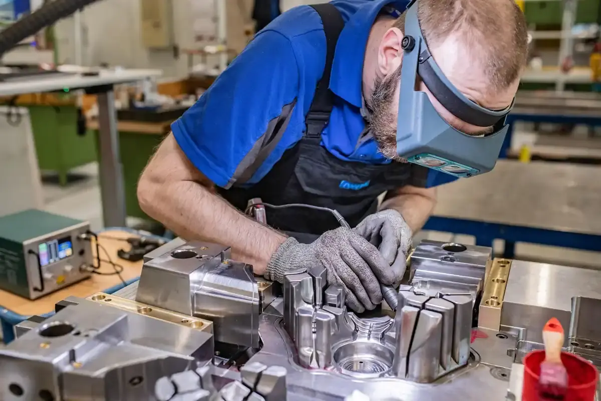 Technician wearing a headband magnifier performing precise fine-machining and repair on a complex metal mold tool in a workshop.