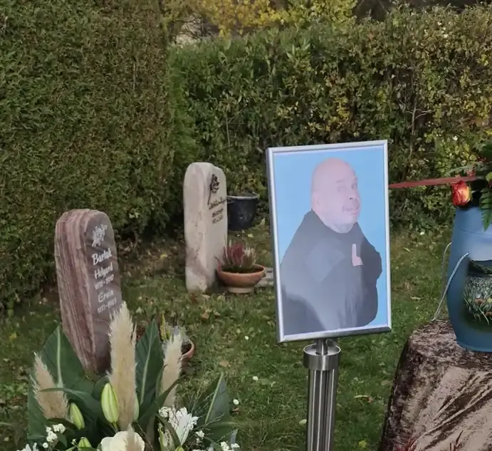 Memorial set-up during the funeral service for an AWM team member, featuring a framed portrait, funeral flowers, and an urn display outdoors.