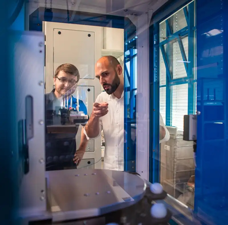 Two AWM employees reviewing and commissioning an automated assembly cell behind a transparent safety glass panel.