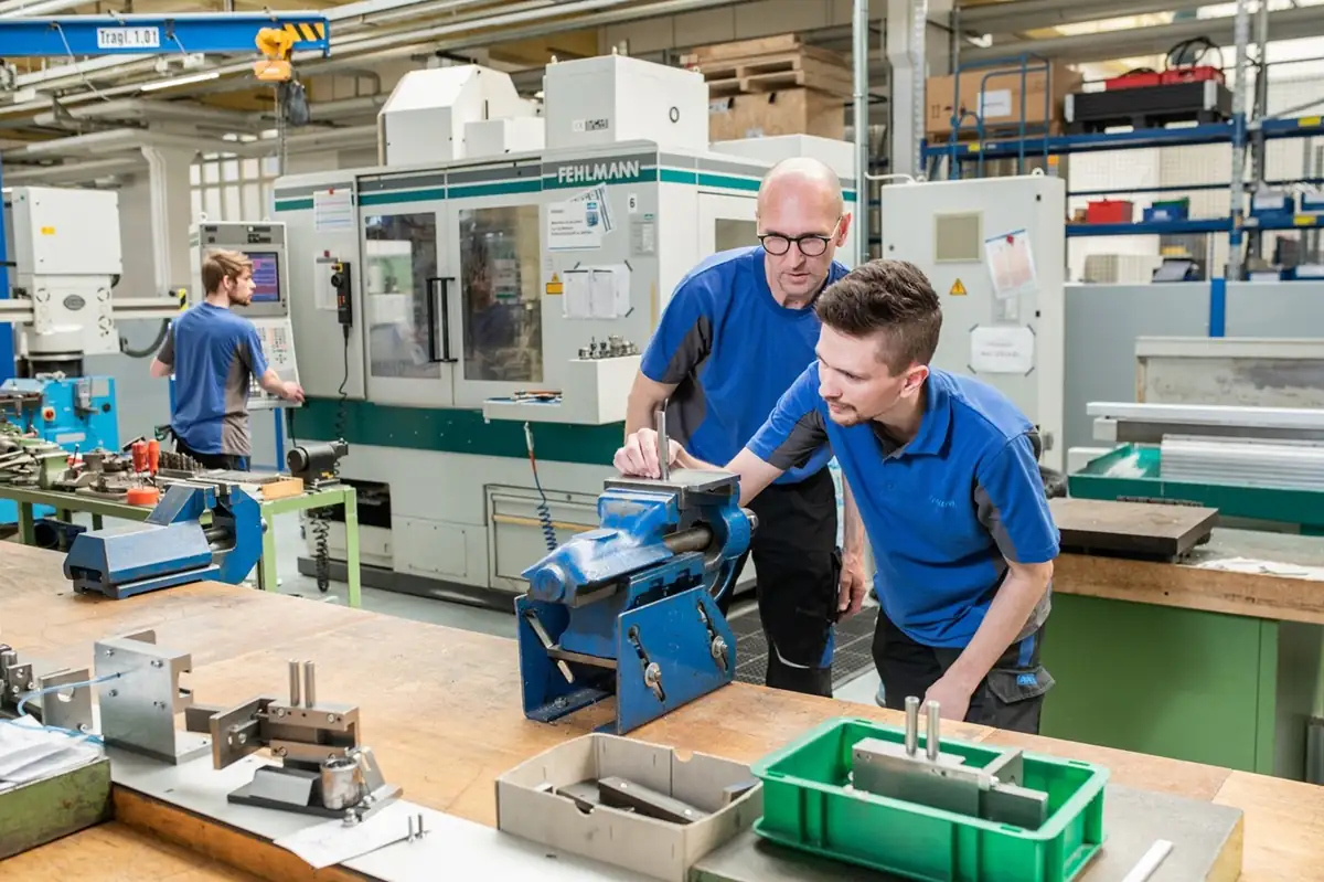 Zwei Techniker prüfen ein Werkstück im Schraubstock auf der Werkbank in einer Fertigungshalle der AWM GmbH mit CNC-Maschinen im Hintergrund.