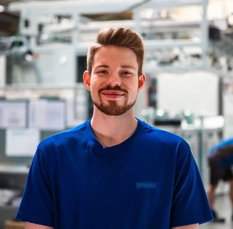 Portrait of a smiling young man wearing a blue AWM work t-shirt in the production hall