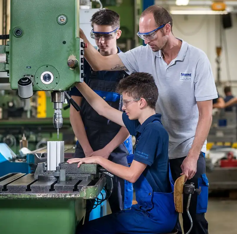 Instructor at AWM Arnstadt guiding two apprentices during precision machining on a machine tool.